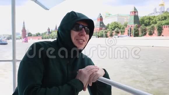戴墨镜的年轻人在乘船旅行时微笑着对着漂浮在莫斯科河上的克里姆林宫的墙壁和塔楼说话视频的预览图