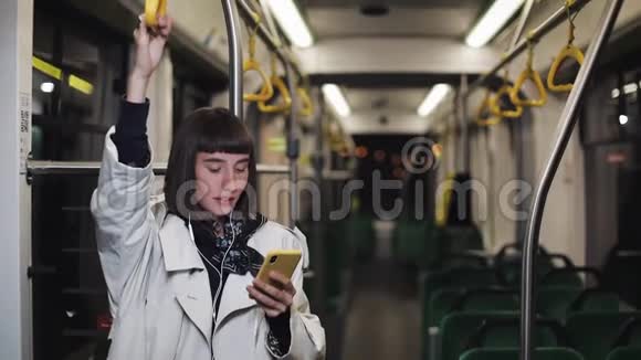 戴着耳机的女士的画像握着扶手听着音乐在公共场合浏览手机视频的预览图