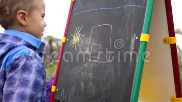 年轻的艺术家在黑板上画画一个男孩在开阔的空气中在鲜花和绿色植物中画孩子们的画视频的预览图