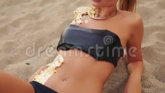 年轻模特穿着黑皮泳衣带着专业的金色艺术妆容躺在海滩上看着相机视频的预览图