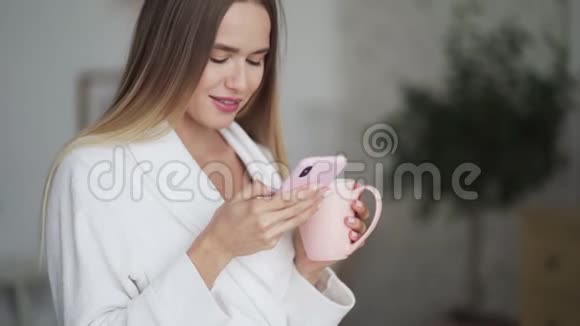 年轻女性的肖像使用电话在厨房端着一杯咖啡或茶视频的预览图