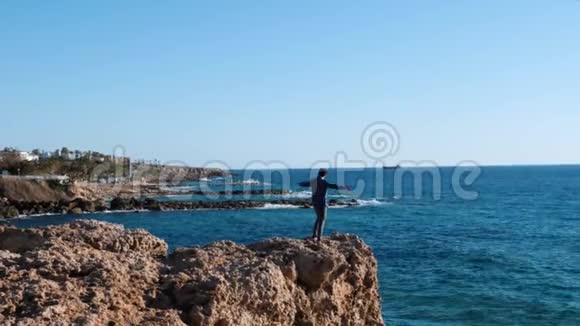 漂亮的白种人穿着蓝色衬衫和牛仔裤站在海边的悬崖边站在岩石上的年轻人视频的预览图