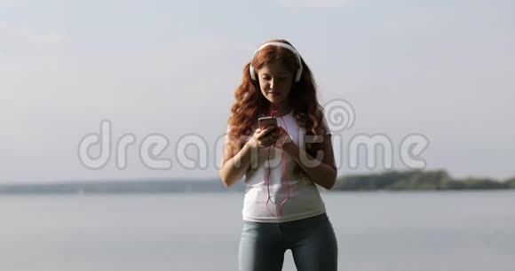 戴着白色耳机的红头发女孩正在手机上选择和听歌视频的预览图