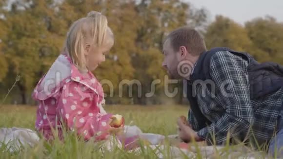 小女孩穿着点缀的粉红色夹克成年白种人坐在草地上吃苹果父亲花钱视频的预览图