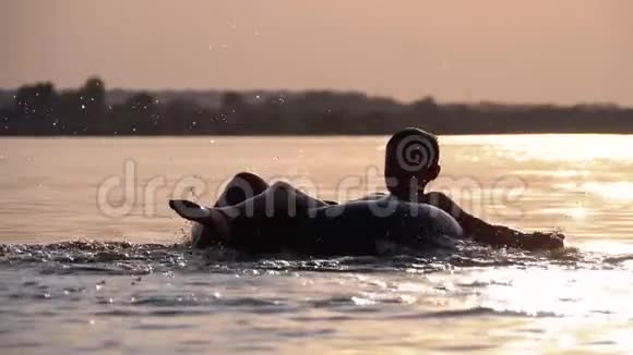 日落时分快乐男孩在河中的充气圈上游泳的剪影慢动作视频的预览图