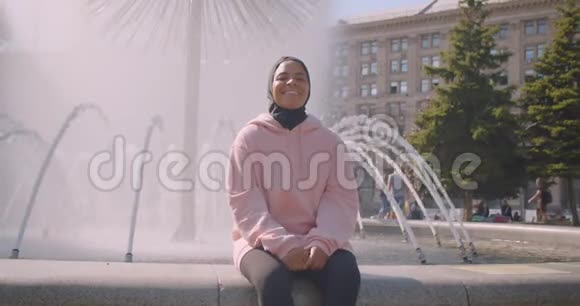 年轻迷人的戴头巾的非裔美国穆斯林女孩的特写照片她开心地坐在上面的喷泉旁视频的预览图
