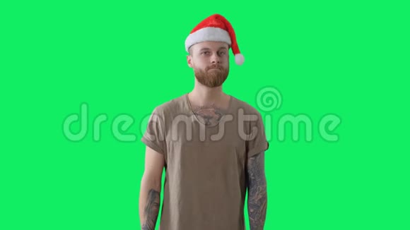 身穿绿色t恤戴着红色圣诞帽长着胡须的年轻纹身男正在彩色钥匙背景上接受礼物视频的预览图