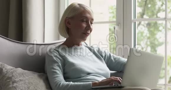 使用笔记本电脑的中年妇女在沙发上看屏幕视频的预览图