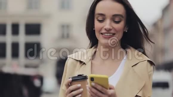 吸引人的年轻女士手持咖啡杯微笑使用她的智能手机和打字走在城市街道关闭视频的预览图