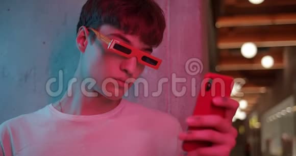 年轻的酷男戴着时尚的现代派对眼镜站在一边靠在墙上用他的智能手机看着屏幕视频的预览图