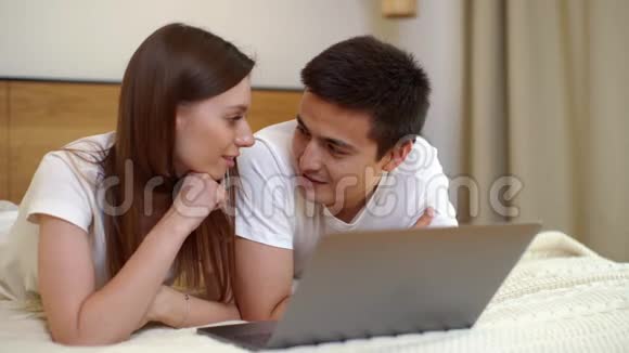年轻的一对女孩和男孩躺在笔记本电脑前的床上在轻型卧室里视频的预览图