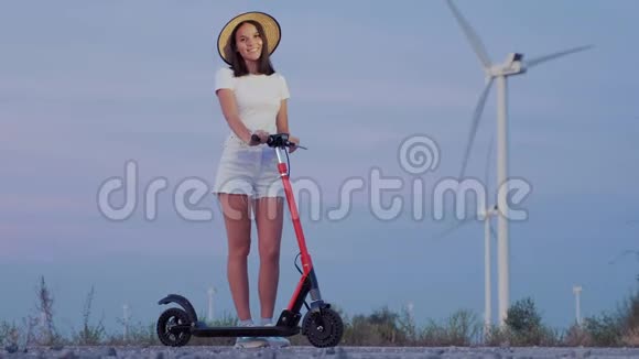 黄昏时分在风力发电机的背景下女孩戴着一顶草帽坐在滑板车上视频的预览图