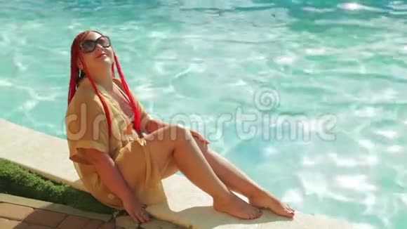 一位穿着浴袍和泳衣的年轻女子正坐在游泳池的边缘视频的预览图