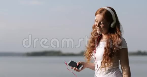 戴着白色耳机的红发女孩正在河边散步和跳舞在手机上听歌视频的预览图