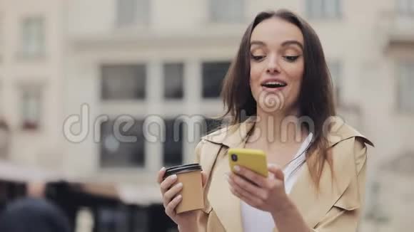 年轻漂亮的女士看着快乐的喝酒用她的智能手机微笑说哇走在城市街上视频的预览图