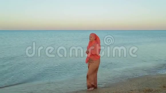 一个穿着橙色披肩的年轻穆斯林女子赤脚走在海滨视频的预览图