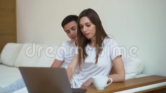 年轻的幸福夫妇在白色柔软的沙发上一起看电影喝咖啡视频的预览图