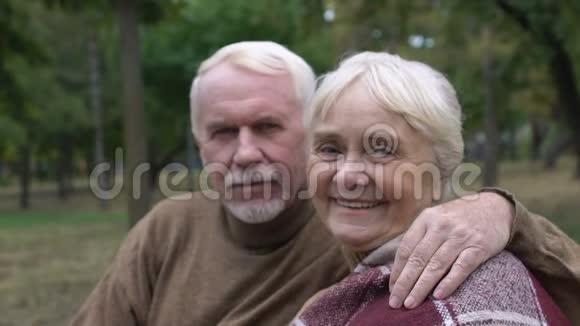 年长的丈夫抱着成熟的妻子坐在公园的长凳上摆姿势拍照视频的预览图