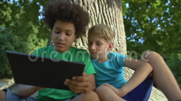 男孩们坐在公园的大树下玩平板电脑朋友们在闲暇时间视频的预览图
