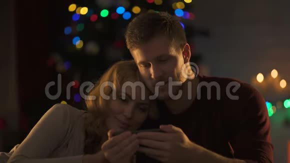 可爱的一对在闪闪发光的圣诞树下在智能手机上看电影亲密视频的预览图