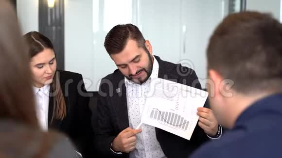 一个英俊的年轻人指着一张图表而他的同事们听着坐在桌子旁视频的预览图