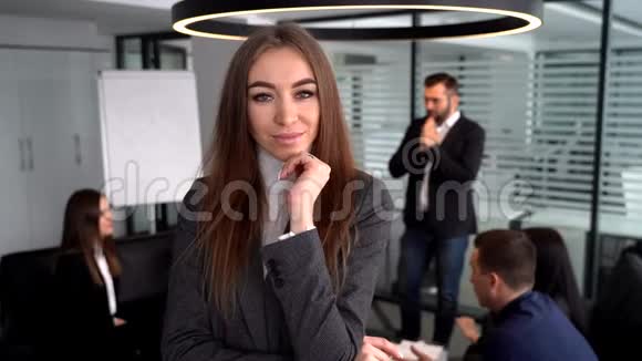 一位身穿灰色西装的年轻女商人为照相机摆姿势同事们在模糊的背景下工作视频的预览图