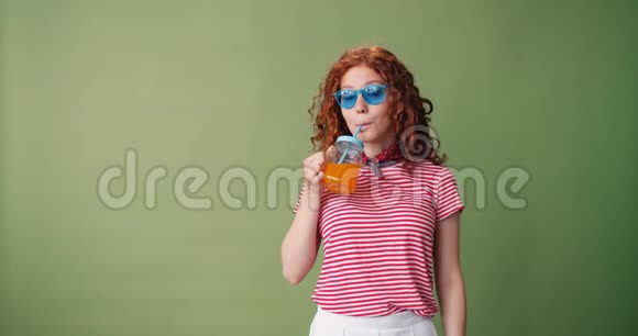 戴着墨镜喝着橙汁在绿色背景下微笑的快乐女孩视频的预览图