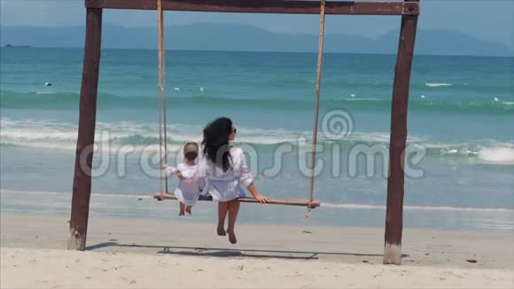 年轻的黑发女孩和她的儿子在海滩上荡秋千暑假玩得开心美丽快乐的女人享受视频的预览图