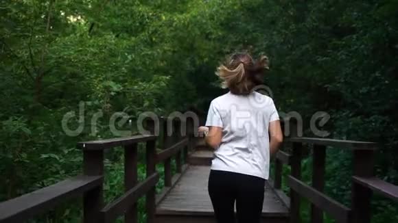 年轻漂亮的女孩或穿着白色T恤和黑色运动裤的女人在公园里跑步回头看她的身后视频的预览图
