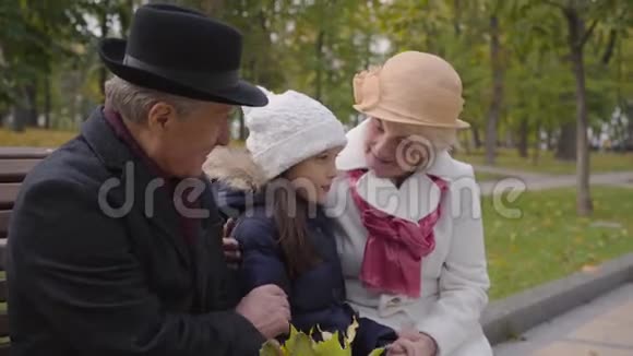 成熟的白人夫妇和他们的孙女坐在秋天公园的长凳上穿着睡衣的祖父母视频的预览图