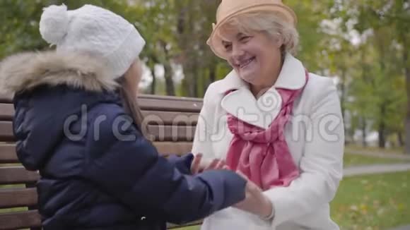 成熟的白种人女人戴着米色的克洛切帽和白色外套和她的孙女坐在长凳上奶奶快乐视频的预览图