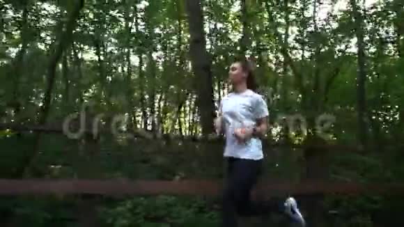 穿着白色t恤和黑色运动裤的年轻运动健美女士或女孩在木桥上的树林里跑步视频的预览图