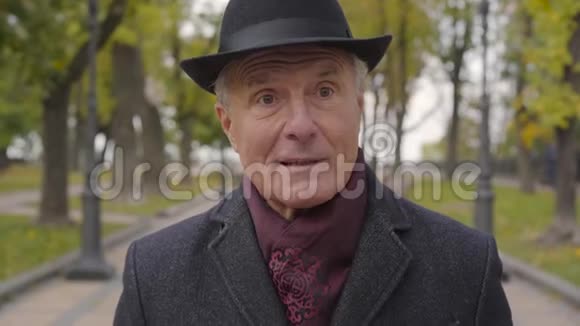 一个成熟的白人男子的特写镜头他的眼睛榛色戴着优雅的黑色帽子和外套站在秋天的公园里视频的预览图