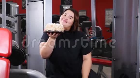 反对运动的食物饮食成瘾的肥胖年轻女子躺在体育馆地板上品尝高热量蛋糕4k拍摄视频的预览图