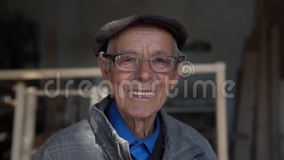 一个戴着蓝色衬衫灰色夹克和帽子的眼镜带着蓝色眼睛看着摄像机的微笑老人闭上了视频的预览图