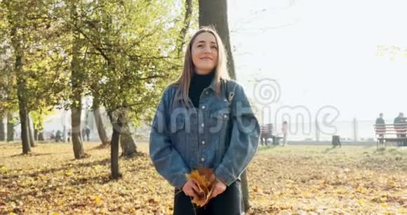 快乐的年轻女人在阳光明媚的秋天公园里扔黄叶慢动作视频的预览图