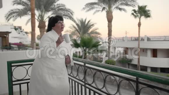 在一个热带度假胜地穿着白色的特里长袍早上在酒店露台上喝咖啡的白人高级妇女视频的预览图