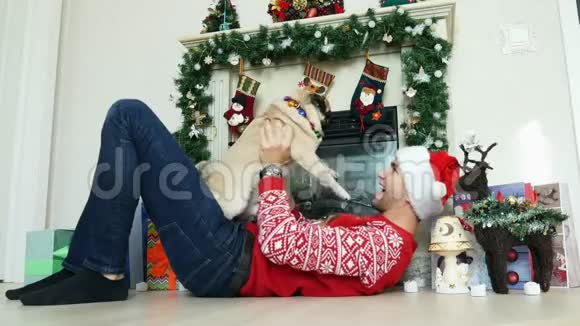 男人躺在圣诞壁炉旁穿着圣诞帽戴着新年和圣诞礼物抱着一只滑稽的小狗视频的预览图