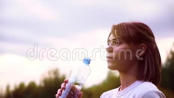 年轻漂亮的运动女孩穿着一件白色T恤很累喝着透明瓶子里的水慢动作视频的预览图