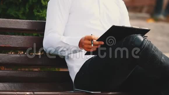 一个年轻的面目全非的商人穿着白衬衫拿着剪贴板和笔坐在长凳上视频的预览图