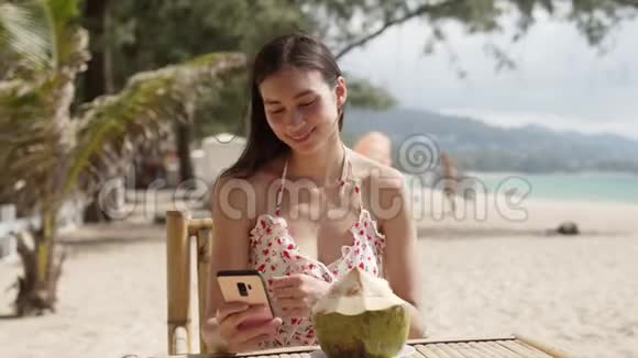 微笑的女人喝椰子水在海滩上使用智能手机视频的预览图