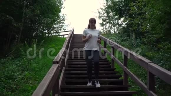年轻漂亮的女孩或穿着白色T恤和黑色运动裤的女人在公园日落时走下楼梯视频的预览图
