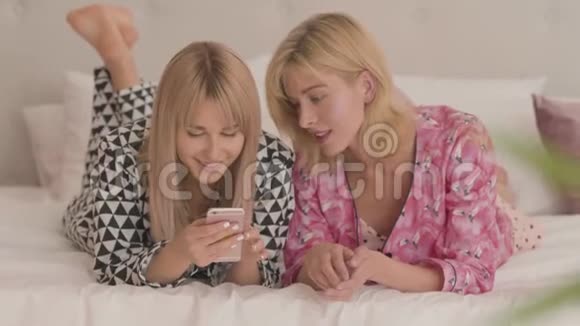 两个年轻的金发白种人女孩穿着睡衣躺在白色柔软的床上看着智能手机快乐的微笑视频的预览图