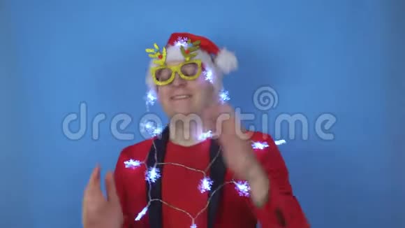 穿着圣诞老人服装和新年灯的年轻白种人正在蓝色背景上跳舞新年假期视频的预览图