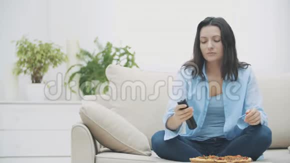 慢点一个年轻的女人正在打开电视节目而坐在沙发上她正在吃面部涂的披萨视频的预览图