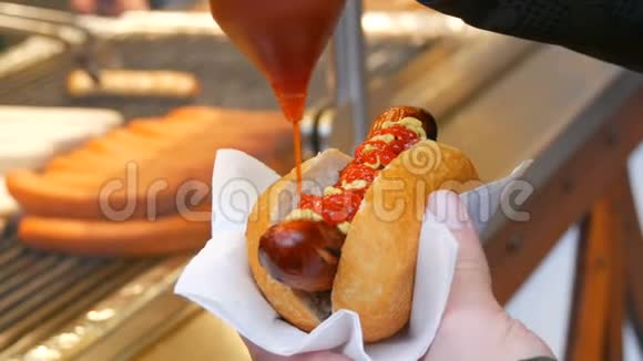 热狗街头食品是不健康的雄性手拿着一根腊肠用番茄酱倒在特制的容器里视频的预览图