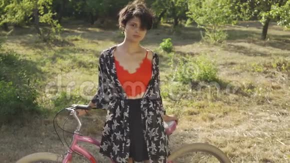 肖像可爱的年轻女子留着短发站在花园或公园里骑着自行车看着相机农村视频的预览图