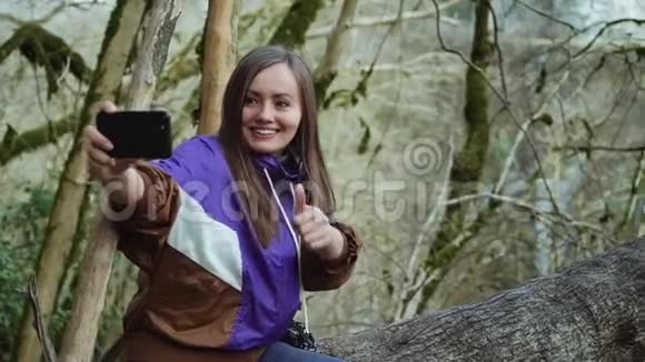 一个在紫色奥林匹亚的快乐女孩在长满青苔的树木的背景下展示自己并制作自拍视频的预览图