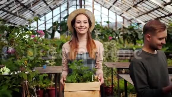 微笑的花店女人在温室里拿着一盒花的慢动作视频的预览图