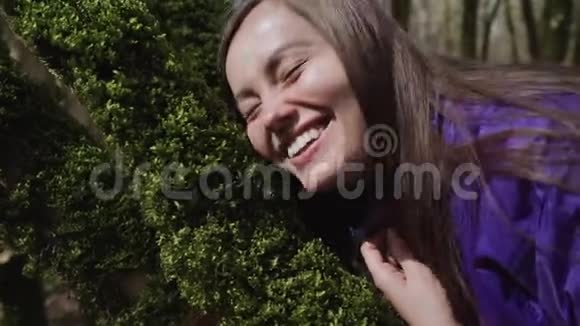 快乐的女孩用她的脸触摸树上柔软的厚厚的绿色苔藓视频的预览图
